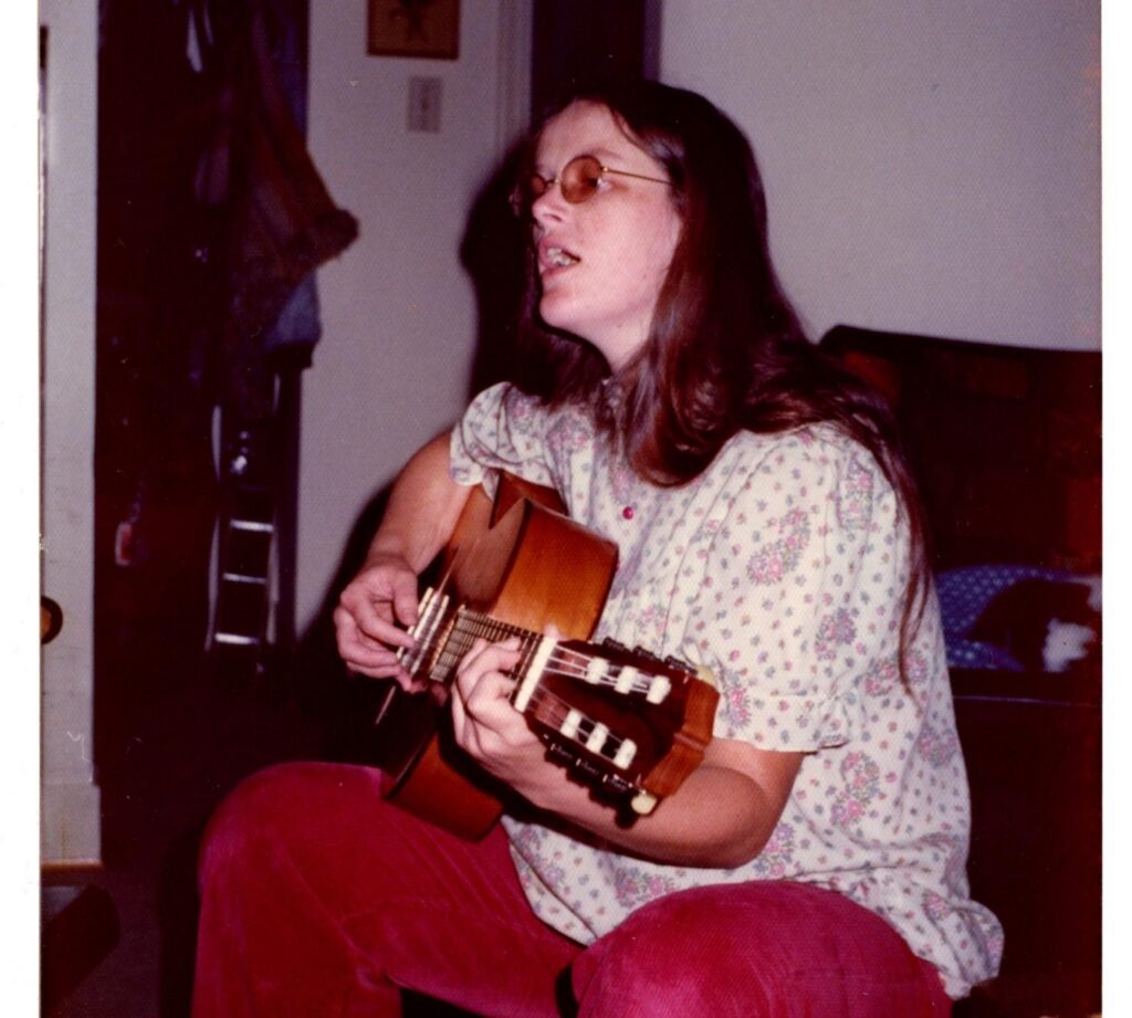 A woman in a white printed blouse and dark orange pants sits on a low seat in a living room, singing and playing guitar. She has long brown hair falling down her shoulders and back, and is pregnant.