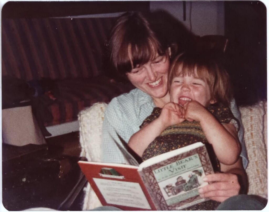 A woman holds a young child on her lap, reading a book. THey are both visibly laughing because they are reading Little Bear's Visit, the child's favourite book.