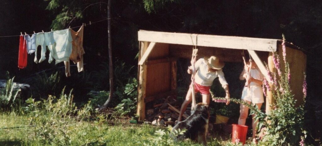 A wild-looking yard with a line full of laundry on the left and a makeshift shed on the right. In the shed, a man and woman stand slaughtering two rabbits, which are hanging from strings at the edge of the shed. The man is handing the kidneys to the couple's black fluffy dog, who is sitting politely, waiting.