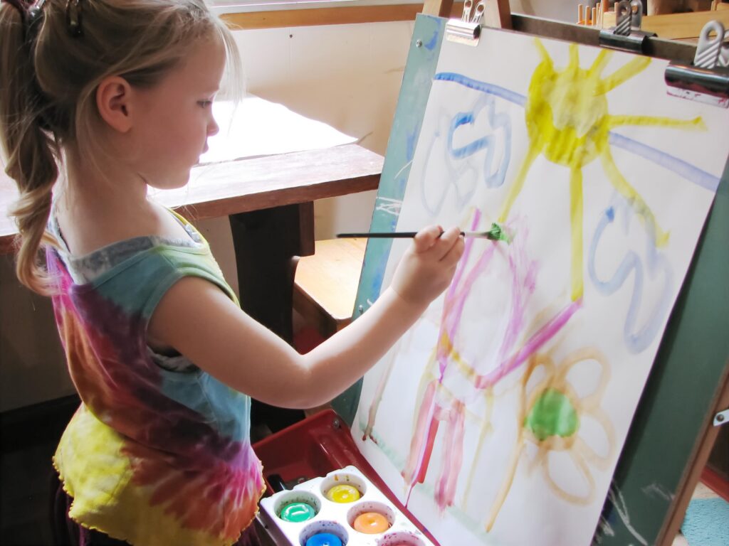 A very young girl with blond hair pulled back into a ponytail, and wearing a rainbow tie-dyed shirt, stands painting at a stand-up easel with a red tray on the bottom, containing a children's cake-paint-palette. She has painted a scene with a blue line of sky, blue clouds, a yellow sun, an orange flower with a green centre, and a big pink person stretching its arms out.