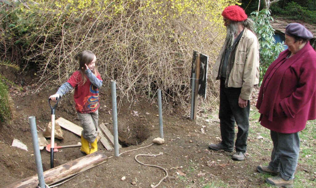 A young boy stands in a small hole in the dirt, holding a shovel in his hand, and surrounded by strewn pieces of wood. A man with a grey beard and long hair stands with a short grey-haired woman, looking at the boy as he describes the "mining work" he is doing.