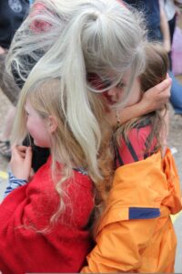 A woman stands bending over her two grandchildren, hugging them. Her long white hair tumbles down over them both. The boy has long brown hair and looks safe and happy under her hair, where she's kissing his head. The girl, with long blond hair, is peeking out from her grandmother's white hair, and smiling.
