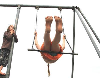 A long-haired boy in a long-sleeved brown shirt and black and white capri pants stands on a bar, half-way up a swing-set, looking wistfully off into the distance. His sister, in an orange skirt, turquoise shorts, and a shirt that is obscured by the angle of the photo, swings on a trapeze, beside him. Her feet stick out into the centre of the photo, in the foreground, and her long braid trails behind her.