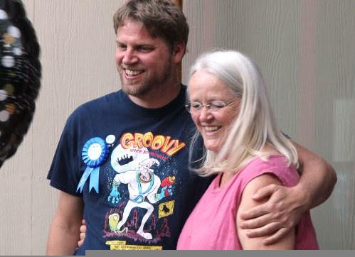 A man with short dark blond hair and a very short beard, wearing a black cartoon t-shirt and a blue ribbon, stands with his arm around his mother. She is shorter than he is, and has long straight white hair, glasses, and is wearing a pink sleeveless shirt. They are both smiling happily.