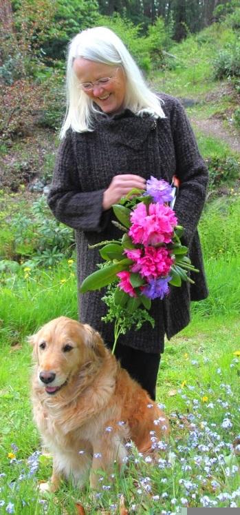 A woman with long straight white hair in a dark brown knee-length sweater and black pants, stands looking down and smiling at a golden retriever, who is also smiling, sitting on her feet, where she stands in her garden. There are forget-me-not flowers all around her feet, and in her hands she is holding a bouquet of raspberry-pink and violet rhododendron flowers, and parsley leaves.