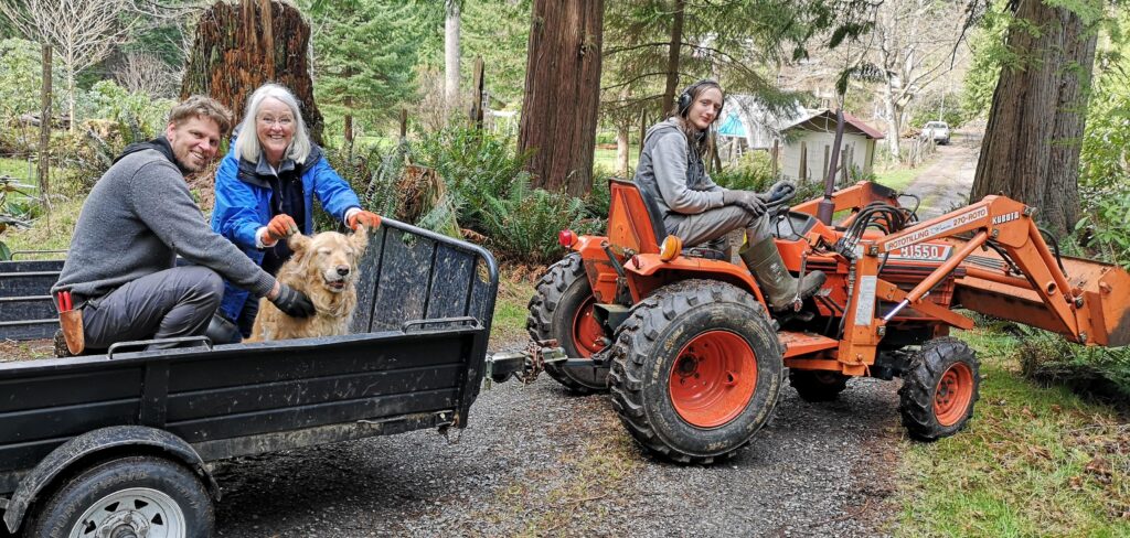 A scene with an orange Kobota tractor hauling a black utility trailer with foot-high sides. A young long-haired man in a grey hoodie is driving the tractor, but turned to the camera, smiling. In the trailer an adult man with short dark blond hair, jeans, and a grey sweater, is smiling, with his hands holding a smiling golden retriever. A woman in a blue rain jacket with long straight white hair stands at the side of the trailer, and is lifting the dog's floppy ears up so it looks like she's flying. This is a happy family portrait on a work day.