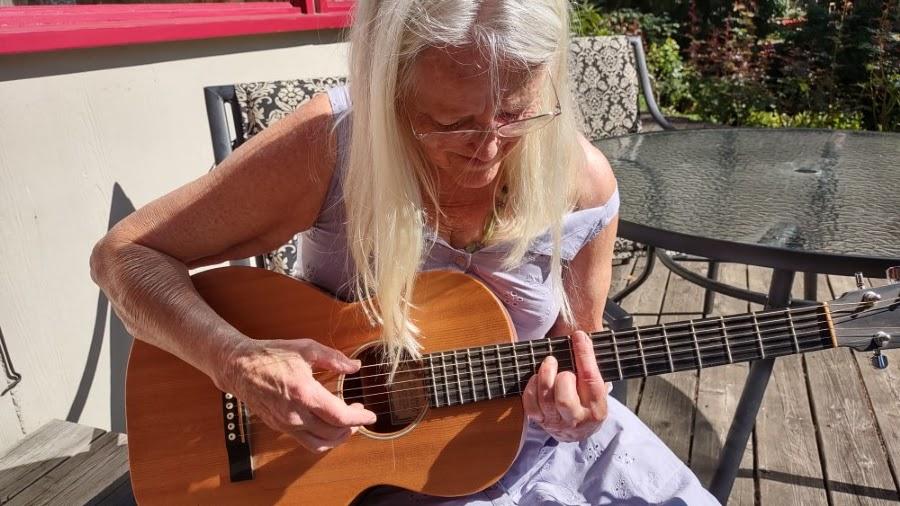 A beautiful woman with long straight white-blond hair sits on a porch, playing a small acoustic guitar. She's looking down at her fingers forming the chord. She's wearing a pale lilac dress and a pair of glasses. It's a sunny day.