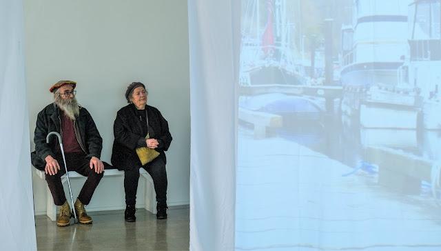 Two artists in black pants and jackets sit on a bench of the Gibsons Public Art Gallery, framed by hanging projections of Emily van Lidth de Jeude's show, what dot home. The man on the left wears a colourful tam, and black-rimmed glasses, and has a long grey beard and hair. His grey walking cane rests between his hiking boots. The woman to his left wears a darker tam, has short grey hair, and holds a yellow purse in her lap. Both are looking thoughtfully at the projections on the hanging sheets, around them.