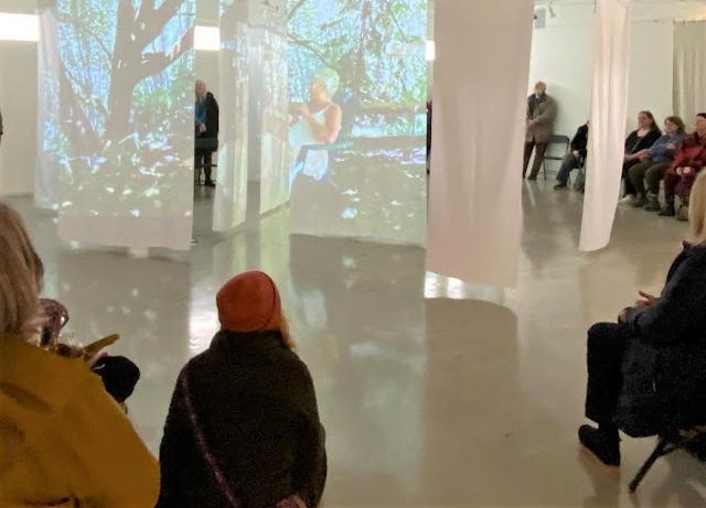 Seven white sheets are hanging in the middle of a gallery with concrete floors, covered with projections of a deciduous forest scene within which a woman sits on a chair ,talking. People are gathered in chairs and seated on the floor all around the exhibition, listening to the show.