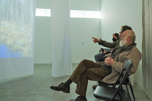 Two men are seated near a gallery wall, smiling pointing at sheets hanging in the middle of the room, which have landscape projections on them. In the distance, another woman is seated, wearing a black respiratory mask.