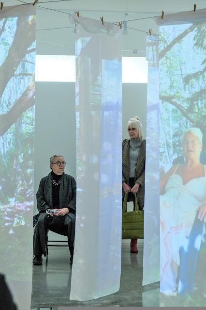 Two artists, Judi Gedye and Diane Buchanan, are framed by separate sections of the hanging white fabric of what dot home. Judi, on the left, is seated on a bench and wearing mostly black with a grey shawl-like coat and a white necklace. She has grey hair and black-rimmed glasses. Diane, standing on the right, wears red boots,a grey turtle neck, a green-grey jacket, and holds a green bag in front of her legs. She has long straight white hair, tied back. On the sheets, a projection of poet Jude Neale is shown. Jude is wearing a white and coloured summer dress, and has short white hair.