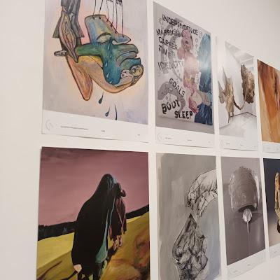 Eight art posters hang on a wall. They're fabulous, but also hard to see because of the angle of the photo and the light reflection on them! Some figurative; some not. It's a great wall of posters by fabulous women, but you'll have to check out the link below it if you want to see more work!!