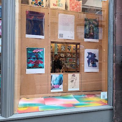A shop window in Scotland shows a series of art posters on tack-board, surrounding a peekaboo window-within-a-window, where a person is seen shopping. One of the posters shows a naked woman sitting on a chair, legs spread, and holding a pair of giant pink plastic lips in front of her crotch. There are rainbow watercolour paintings on the horizontal surface at the bottom of the window. Yay rainbows!!!