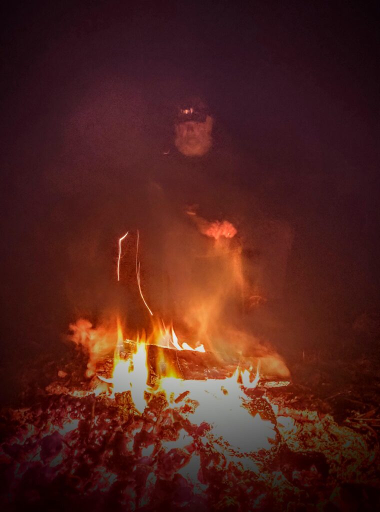 A large fire burns in the foreground, and behind it, semi-obscured by flames, a man with a white beard and baseball hat sits, looking into the flames. he looks sad, and is holding his hands together in front of his knees.