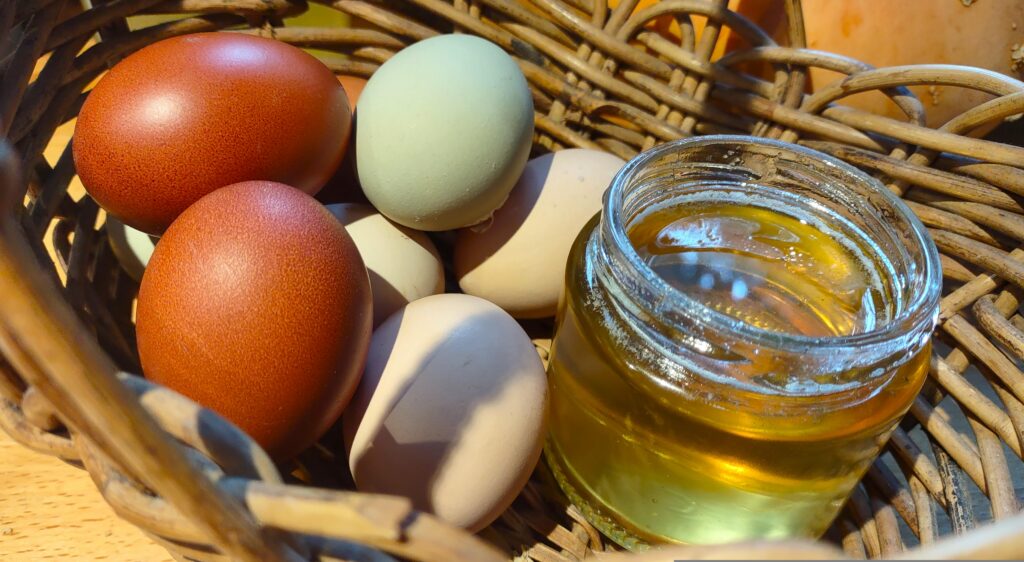 A wicker basket containing nine eggs of different colours (dark reddish-brown, pale brown, green, and pale green) along with a small open jar of local honey.