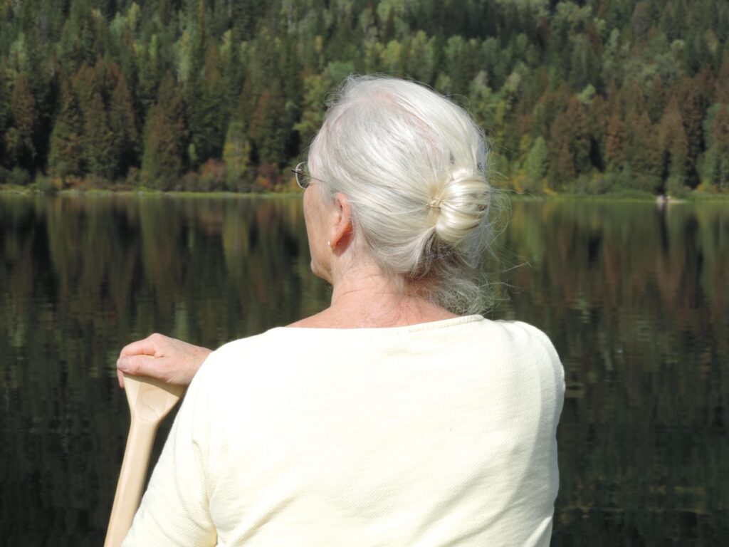 A woman sits at the front of a canoe, paddling. She has long white hair pulled back into a bun, and is paddling on her left side. The man who took the photo, from the back of the canoe, was her husband and the love of her life.