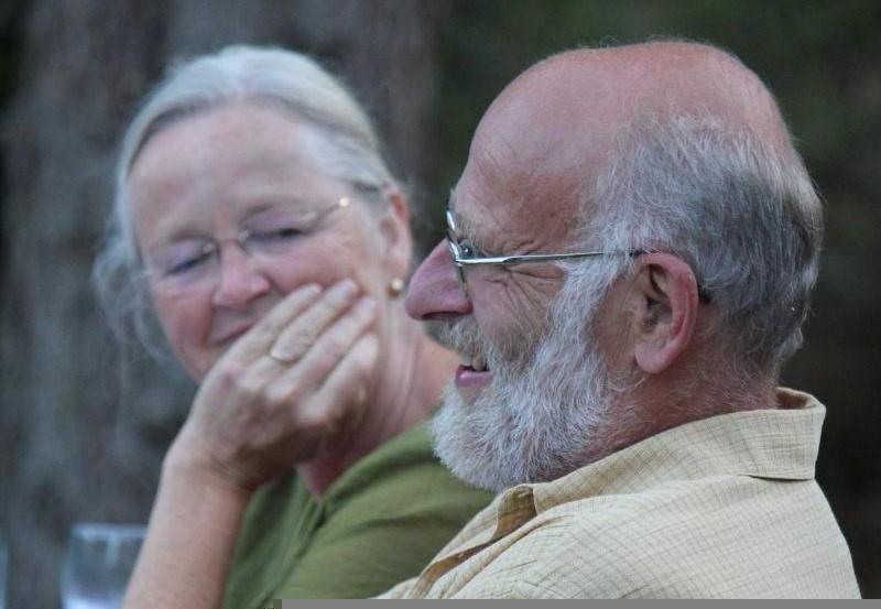 A man with a short grey and white beard and glasses sits smiling in a pale yellow collared shirt, looking to the left. A woman with long white hair pulled back looks at him with her hand held partially in front of her face. She is smiling and really her whole face beams with love and affection for this man, who is her husband.