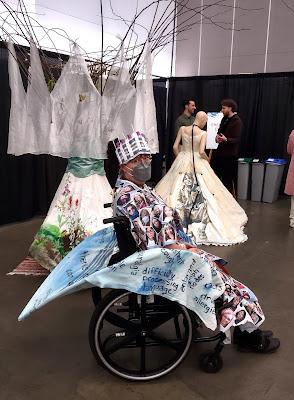 The artist, Emily van Lidth de Jeude, sits in her wheelchair, wearing the Hospital Gown project she created. Hospital Gown is a wearable art piece covered with selfies of over 300 Long Covid patients, and a train (which she's sitting on) covered with many of the most common symptoms of Long Covid. In the distance, two more of Emily's wearable art pieces can be seen, on display.