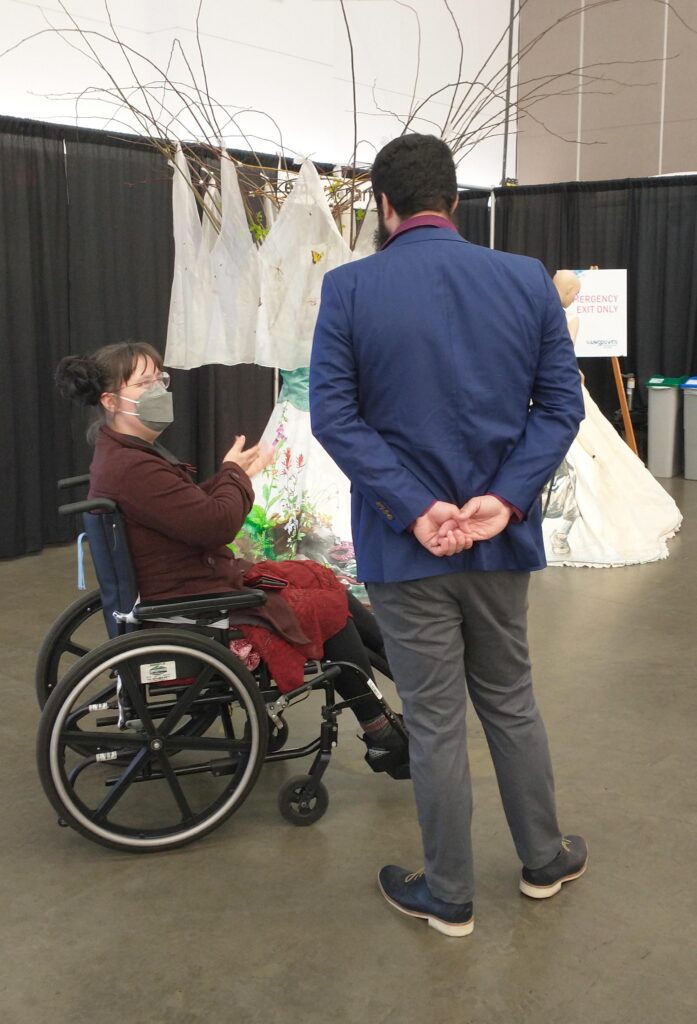 Artist Emily van Lidth de Jeude sits in her wheelchair, wearing black leggings, a short red dress, a burgundy jacket, and a grey face mask. She has two curly fake hair buns, and is gesturing with her hands as she speaks to a dark-bearded man in grey pants and a blue suit jacket. Some of Emily's wearable art pieces are visible in the background.