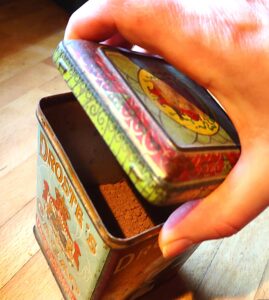 A hand lifts the lid of an antique Droste's Cocoa tin. There is red dutch-processed cocoa powder, inside.