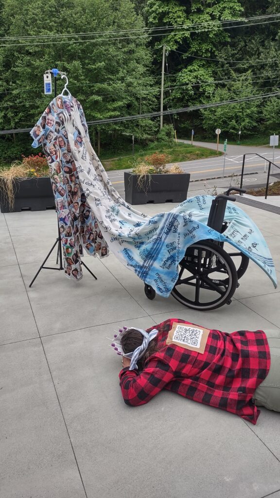 A different view of the blue hospital gown covered with the faces of over 300 Long Covid patients, hanging from an IV pole, standing on the concrete patio of the Bowen Island Community Centre, with planter boxes and a road, behind it. The back of the gown is now visible, where a long train made of a hospital blanket is attached to and covering a wheelchair. The train is covered with the names of common symptoms of Long Covid, and is attached to the back of the gown with a pair of white fabric hands. Artist Emily van Lidth de Jeude is lying on the ground, wearing a red lumber jacket, green pants, and with a QR code pinned to her back. She is wearing a crown made of hospital blanket, blood vials, and covid testing kits.