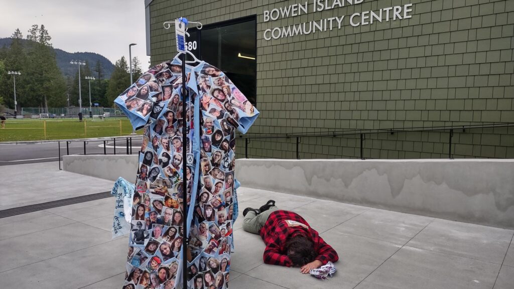 A photo of a blue hospital gown covered with the faces of over 300 Long Covid patients, hanging from an IV pole, standing on the concrete patio outside a building that says Bowen Island Community Centre. Artist Emily van Lidth de Jeude is lying on the ground, wearing a red lumber jacket, green pants, and with a QR code pinned to her back.