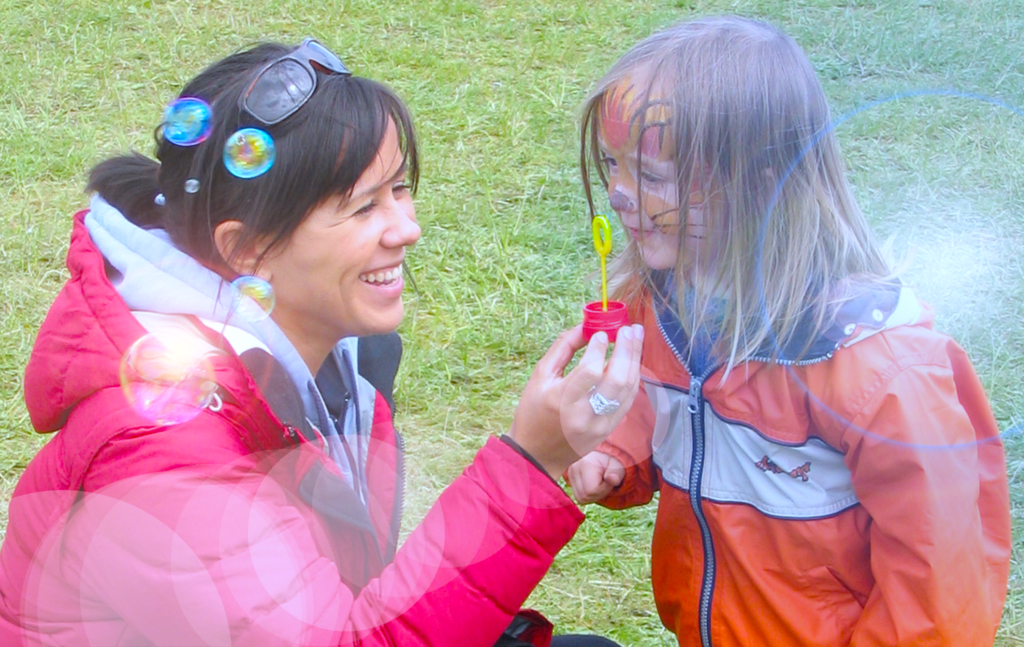 A young boy with long blond hair and his face painted like a tiger blows rainbow bubbles through a bubble wand that's being held by a smiling Hispanic woman who has long black hair.