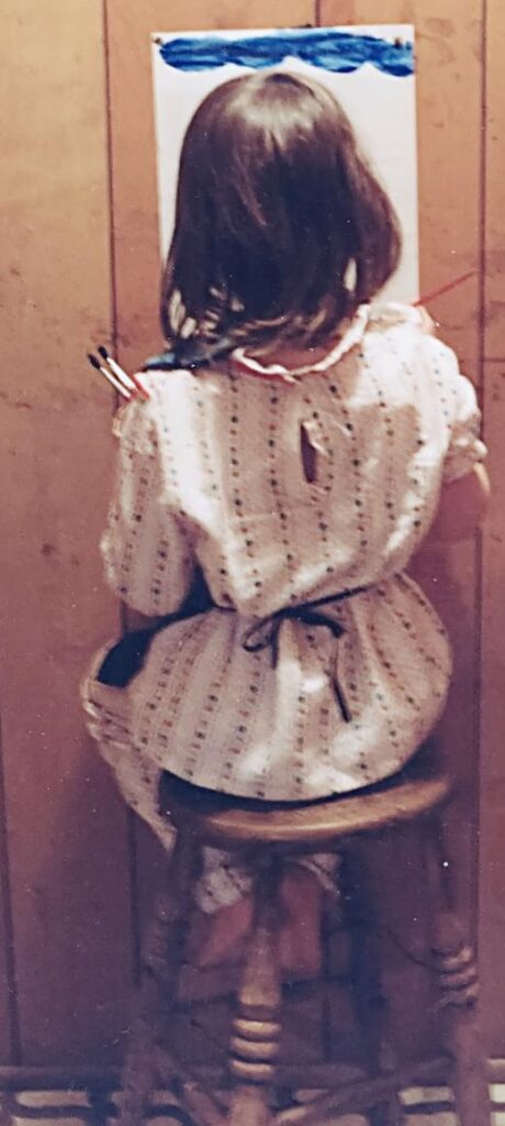 A young girl with straight brown hair in a white nightie and apron is seen from behind, seated on a wooden stool, facing a wood-panel wall. She is painting on a piece of paper that's taxed to the wall. She has painted blue clouds at the top of the page, and holds brushes in her free hand, on the left.