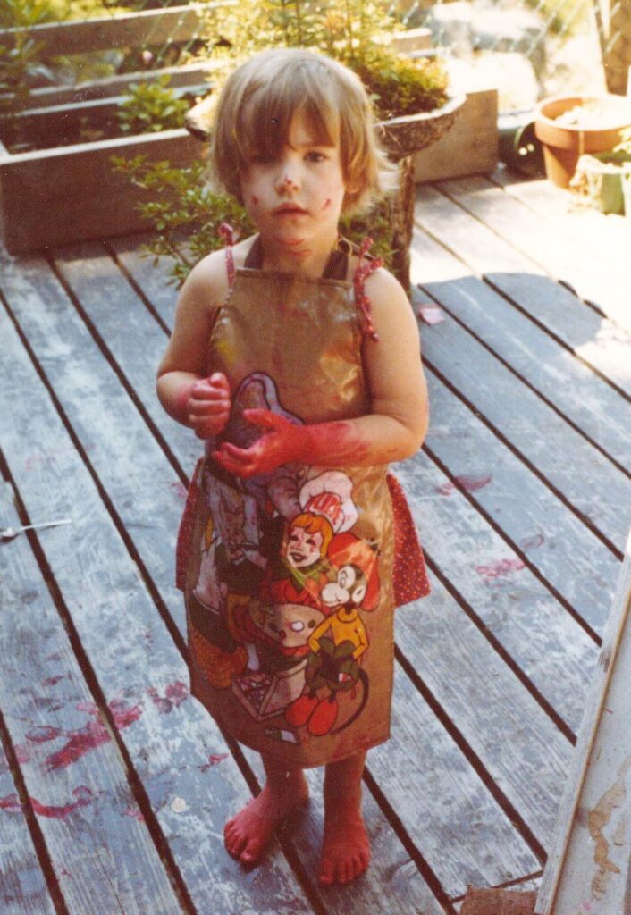 A child in a plastic cartoon apron stands outside a sliding door on a wooden porch. She has short straight brown hair, and her hands, feet, and part of her face are completely covered in red paint. There are also red footprints all over the porch.