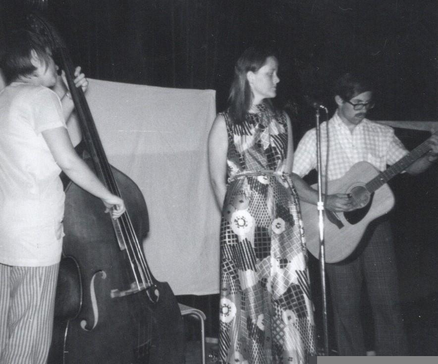 A black and white photo of a band on stage. Woman on left plays bass violin, woman in the middle is Lyn and stands not singing at a microphone, waiting as the man on the right, with a large acoustic guitar, appears to be taking a solo. Lyn is wearing a loud printed dress, 60's style.