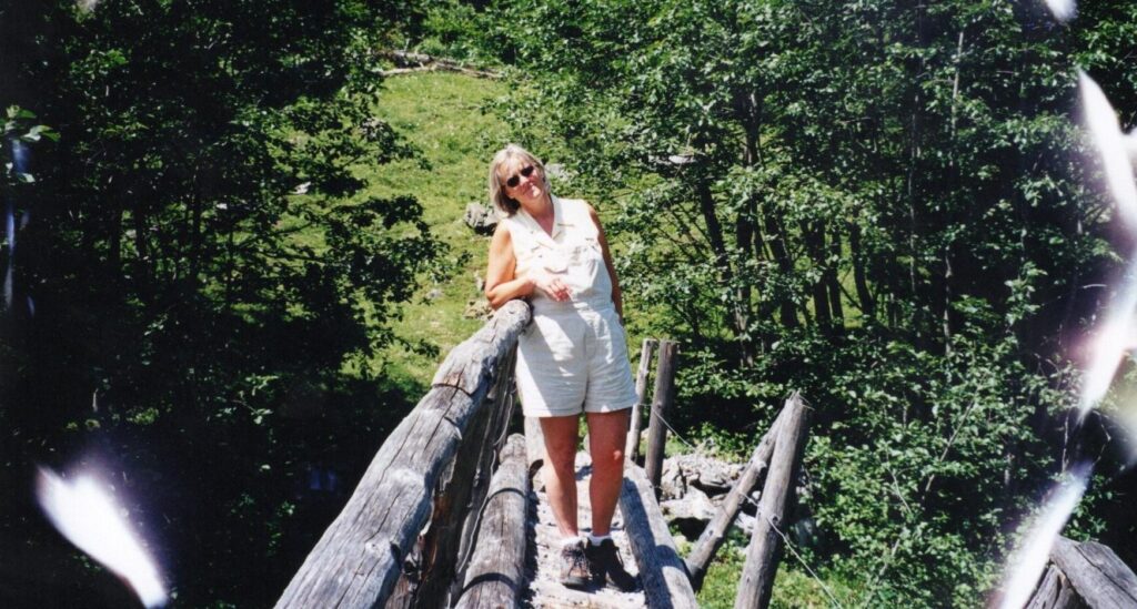 A woman with short grey and white hair stands on a handmade footbridge in the Swiss alps, smiling at the camera because the love of her life is behind it.