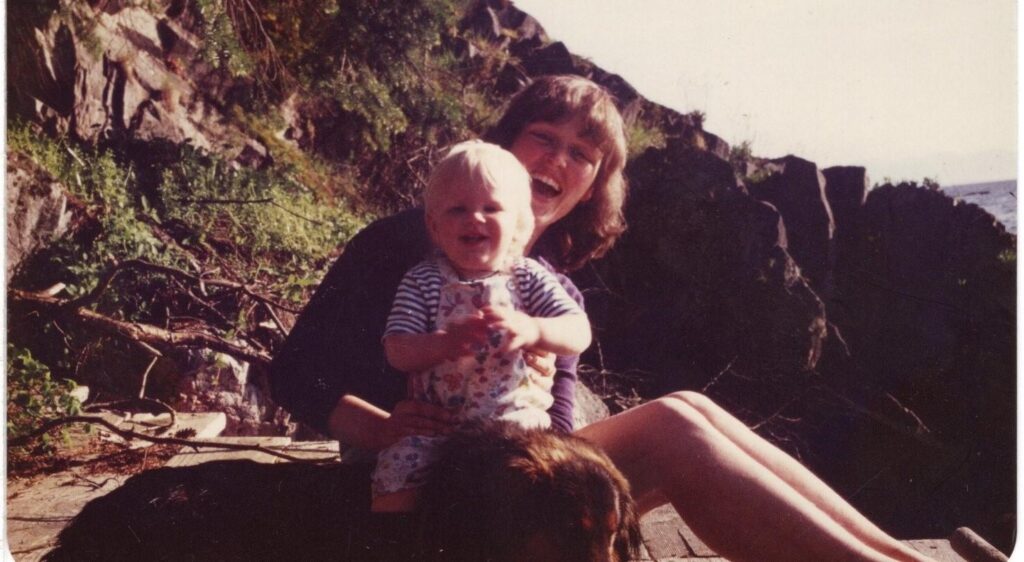 A woman sits on a dock with seaside bluffs in the background. She is laughing at the camera, and holding a little laughing boy ontop of their big fluffy dog.