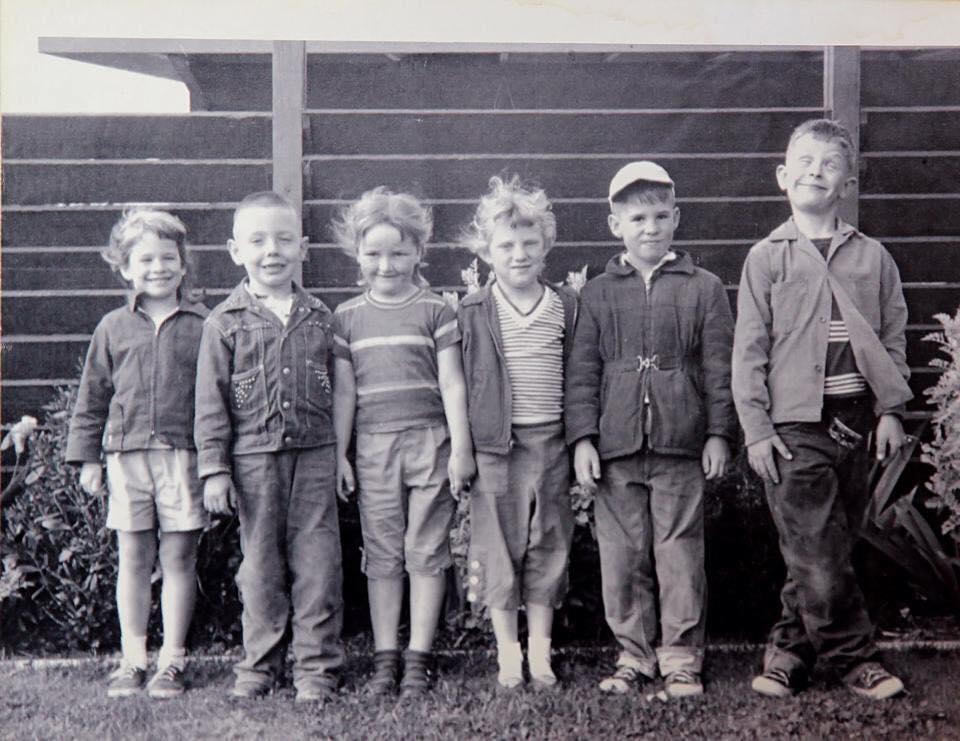 A row of six children stands against a few shrubs and a wooden fence.