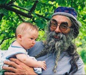 A close-up photo of a man holding a baby under an apple tree. The man has long grey and black hair and a braided beard. He is wearing a purple hat and a blue T-shirt, and holding the baby who is wearing a white summer shirt with pale blue trim.