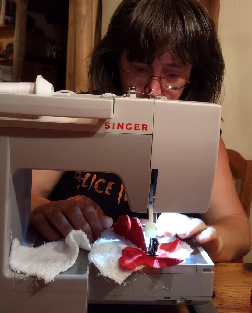 Artist Emily van Lidth de Jeude, seated at her sewing machine, wearing an Alice In Chains t-shirt, sewing a rooster head out of white terrycloth and red felt.