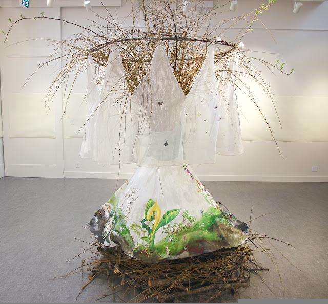 In a white-walled gallery, a wedding dress stands upright above a huge nest of branches, and a massive spray of willow branches emerges out the top, where a body and head might otherwise be. The skirt is painted with a landscape of Pacific Northwest seasons around. Visible in this image is spring: A damp mossy hummock with trillium, skunk cabbage, fiddleheads, and various small blooming wildflowers. From the top of the huge spray of willow branches, a circular four-foot-wide veil is hanging all the way around. The veil is painted with flying insects, through the seasons. The view in this photo is sprint, and the insects include various northwest coast species of mosquitoes, bees, beetles, butterflies flying ants, and moths.