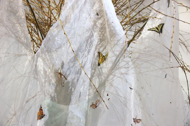 A close-up view of the disrobe gaia gown's insect-veil. Visible in this view is a small section of the veil, hanging down from a spray of willow branches. On the veil we can see a lot of flying insects: butterflies, a june beetle, moths, a crane-fly, mosquitoes, and flying ants.