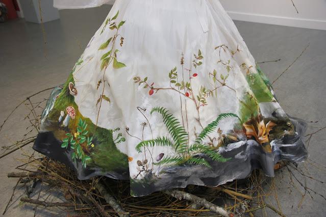 A white wedding dress skirt is shown, painted with plants of the pacific northwest as they might appear in late autumn and early winter. A tall dying stinging nettle laden with seeds is on the left, with amanita mushrooms, a bracket fungus, mosses, licorice fern fiddleheads, a browning sword fern, dried maple leaves, dried aster seedheads, and a baldhip rose with a few red rosehips on it.