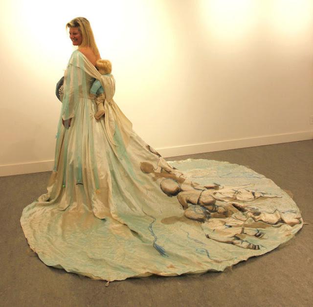 A woman stands with her back turned mostly towards the camera, looking over her shoulder and smiling. A pregnant belly protrudes from the dress and is painted but we can't see with what in this photo. The dress's enormous train is painted with seven babies clambering up to the person wearing the dress. At the back of the dress, there is a built-in baby-carrier, with a large baby doll hanging in it. The dress has soothers poking out of the bodice where the wearer's own nipples would be, and blue ribbons hanging from the shoulders, with more soothers on the ends.