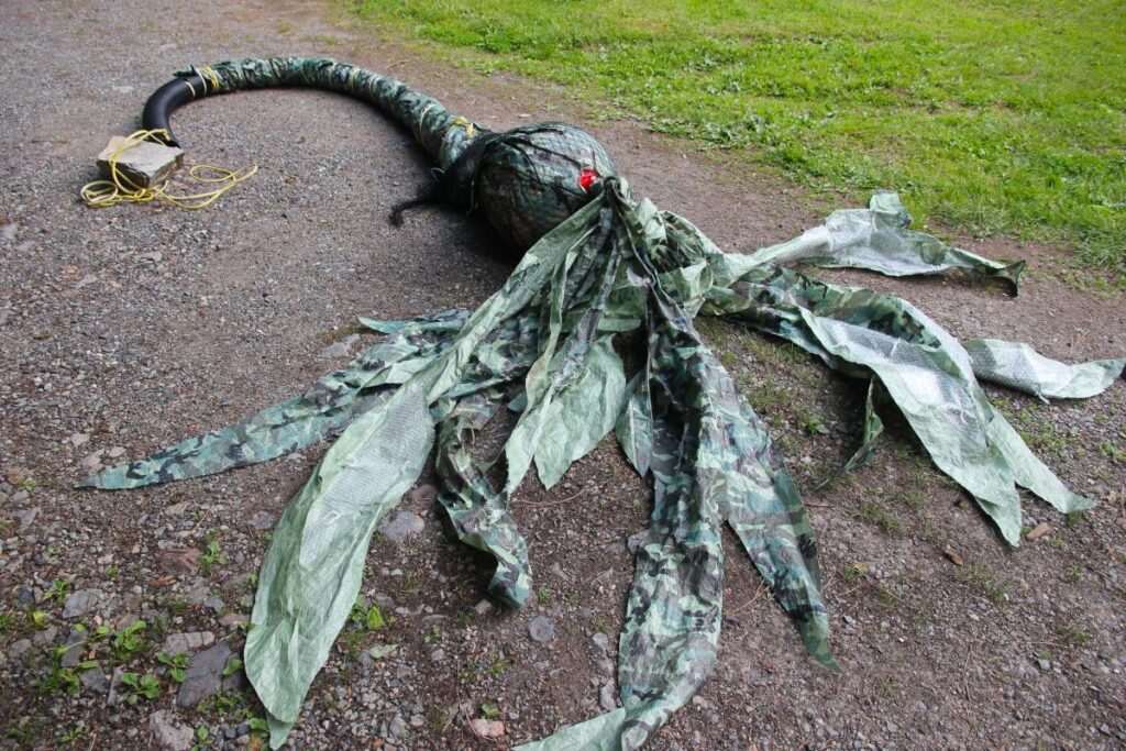 A photo of a giant bull-kelp made of garbage, lying on a rocky ground, with bits of grass at the edges. The bulbous top of the kelp is nearly two feet wide, and a large volume of plastic leaves spread out on ground around it. The whole thing is created from a 12-foot-long flexible plastic drainage pipe, a marine buoy, a green and black camouflage tarp, and various plastic ropes. 
Artist: Emily van Lidth de Jeude