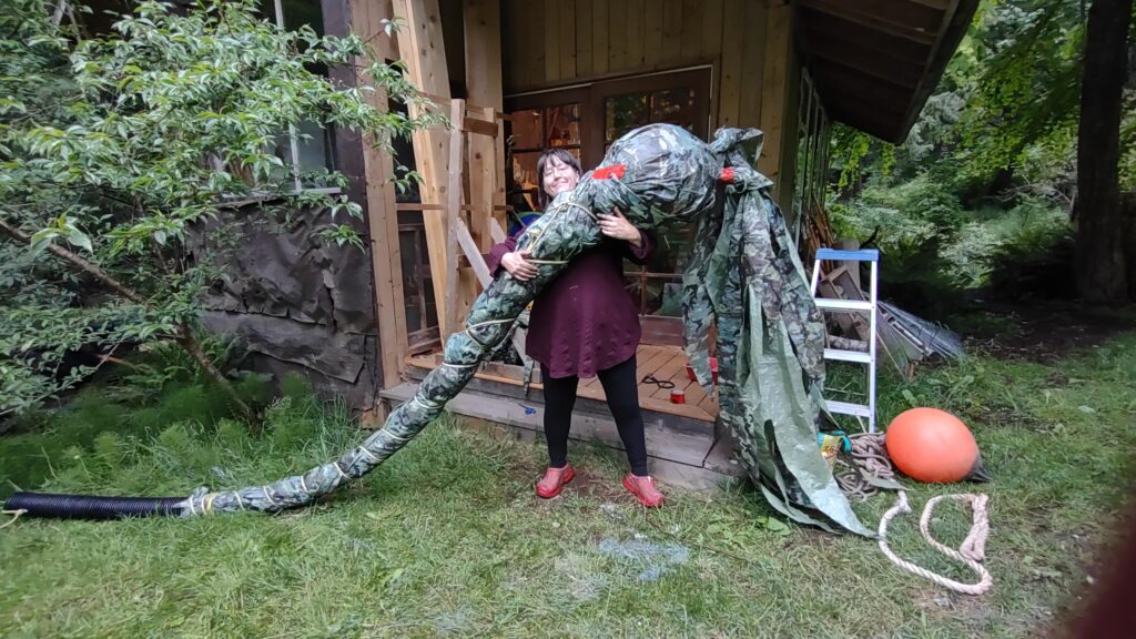 Artist Emily van Lidth de Jeude stands in front of her wood-walled studio, on a lawn, holding up a giant bull-kelp made of garbage. The bulbous top of the kelp is nearly two feet wide, and a large volume of plastic leaves hangs off the end of it. The whole thing is created from a 12-foot-long flexible plastic drainage pipe, a marine buoy, a green and black camouflage tarp, and various plastic ropes. 
In the background and around Emily are some remains of her work: a roll of red tuck tape, some ropes, shreds of cut plastic tarp, a smaller buoy, a step-ladder, and various pieces of lumber. On the left side of the photo is also a young peach tree, growing against the wall of the studio.