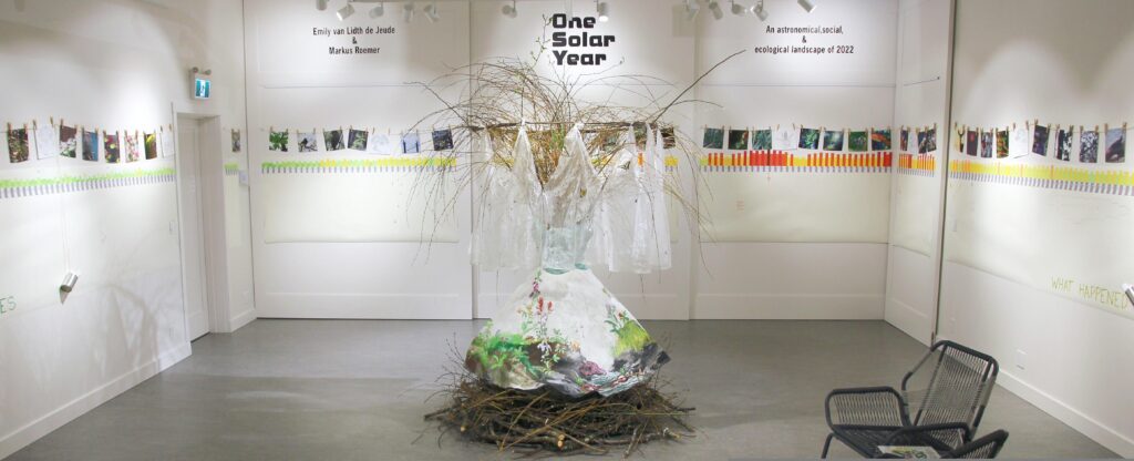 A wide view of a gallery with an installation of the cycle of seasons. From left to right, the gallery walls are covered in a graph showing daily and nightly temperatures, along with solar, lunar, and tidal cycles, and weather observations. Above the graph are photo prints hung by clothespins, depicting natural features like plants and small animals, that correspond with the days in the cycles graph. Under the graph is a long white paper, and tins full of coloured pens. This will be used to create a community mural over the course of the exhibition. In the centre of the gallery is a white dress painted with plants and invertebrates of all seasons, and a circular veil, covered with flying insects, also according to season. The dress is displayed on a giant nest of sticks, with a huge bouquet of willow branches, extending out the top. The branches will leaf out during the exhibition.