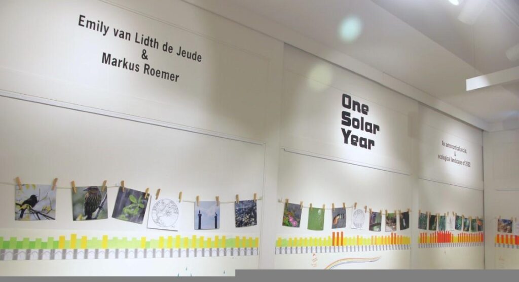 A view of a gallery wall with part of an installation of the cycle of seasons. From left to right, this view shows a graph of daily and nightly temperatures from April through June, along with solar, lunar, and tidal cycles, and weather observations. Above the graph are photo prints hung by clothespins, depicting natural features like plants and small animals, that correspond with the days in the cycles graph.
High up on the wall of the gallery, vinyl text says the title of the show, "One Solar Year", and the names of the artists, Emily van Lidth de Jeude and Markus Roemer.