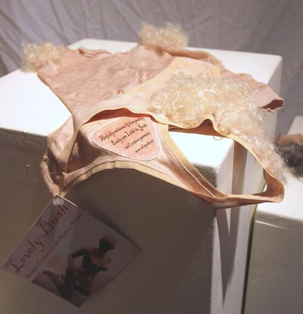 Displayed on a white plinth, the Lovely Bush blond pubic hair set: a pale beige thong covered with very curly blond curls in the front, and a nylon top with very curly blond curls at the armpits. A tag hangs off the side of the plinth that says Lovely Bush: Aesthetic Prosthetics for the Aspiring Woman.
