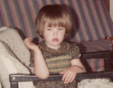 A young girl with short straight brown hair sits in the side of an armchair, wearing a smocked dress covered in small yellow and green flowers. She holds the arm of the chair with one hand, and the other hand hangs in the air beside her face. She's looking down and to the right with a lost, sullen expression, her mouth slightly open.