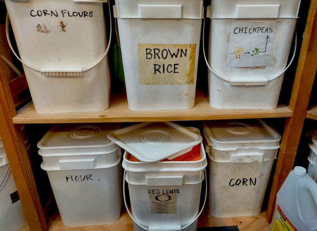 Six large plastic buckets sitting on the floor and a shelf directly above. They have labels on them written in sharpie pen that say: Corn flours, brown rice, chickpeas, flour, rend lentil, and corn. The red lentil bucket is sitting open and red lentils can be seen inside, nearly filling the bucket. A large plastic jug of white vinegar sits to the right of the buckets, on the plywood floor.