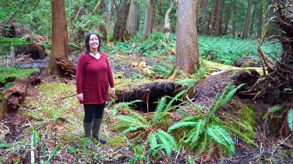 A woman with brown hair and glasses stands in a mossy hemlock and cedar forest. She's wearing gumboots, and there are many ferns around her, and a very large swampy puddle behind her.
