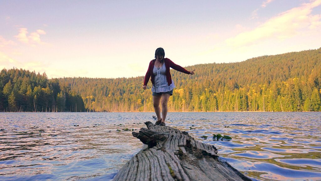 A photo of a woman with brown hair, balancing out on a log in the middle of a lake. There are lily pads in the water beside her, and sunlit hillsides covered with coniferous forest, in the distance.